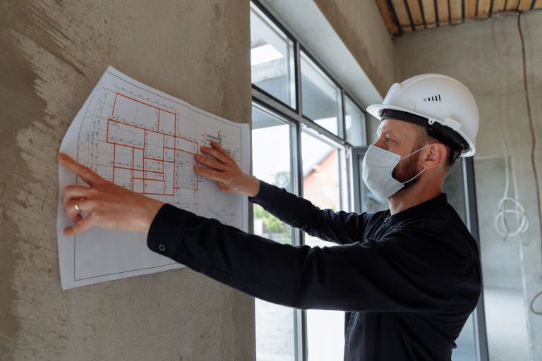 Mężczyzna w kasku patrzy na rysunek techniczny oparty o ścianę wnętrza budowanego domu.